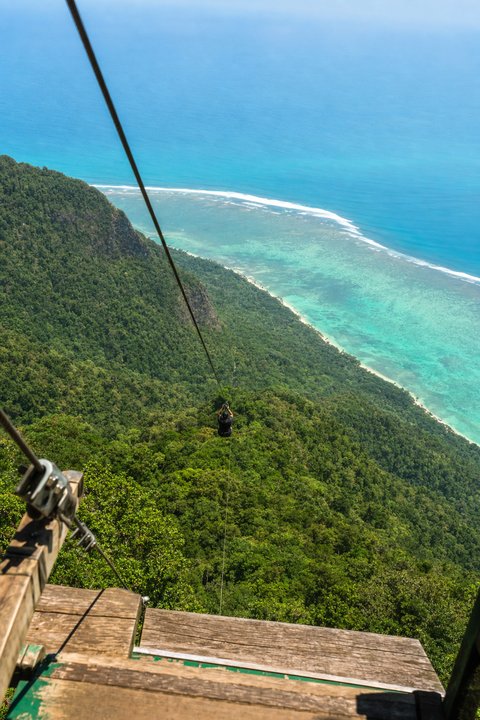 The view down a zipline cable from a launch platform, over steep forested ridge to a turquoise reef coast