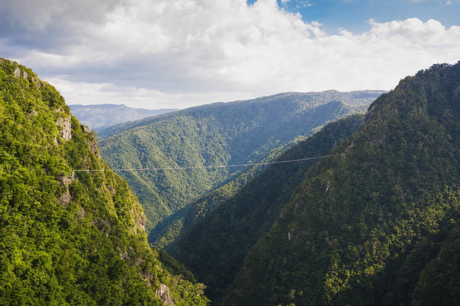 A single very long zipline cable spanning a deep green valley between two steep mountain ridges