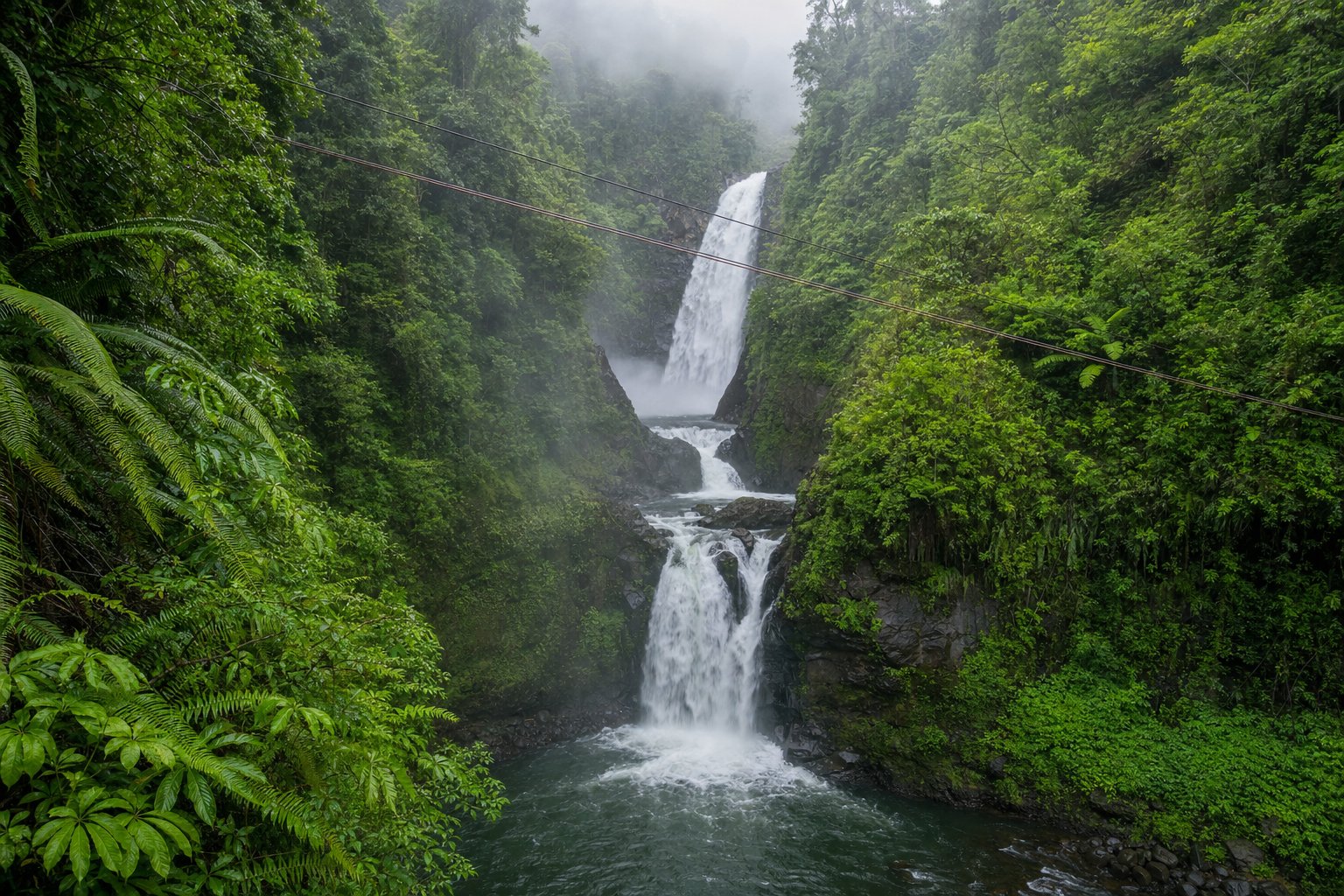 Zipline cables strung across a lush river gorge with tiered waterfalls dropping into a pool