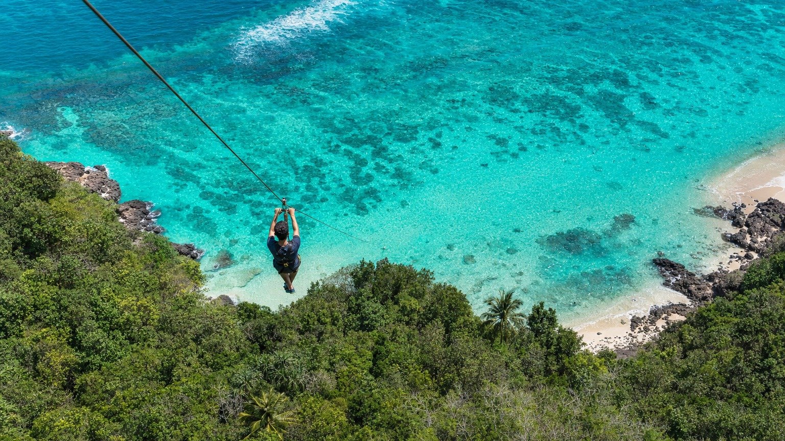 A zipline rider glides on a steel cable above a turquoise reef lagoon and forested coastline